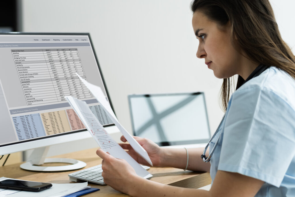 Eine Person aus dem Pflegedienst sitzt vor einem PC mit Patientenakten in der Hand